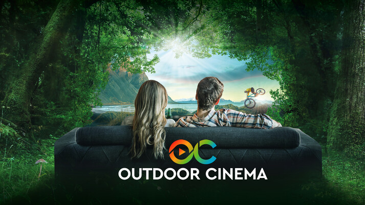 A woman and a man are shown from behind, sitting on a sofa and looking out over a forest and mountain landscape