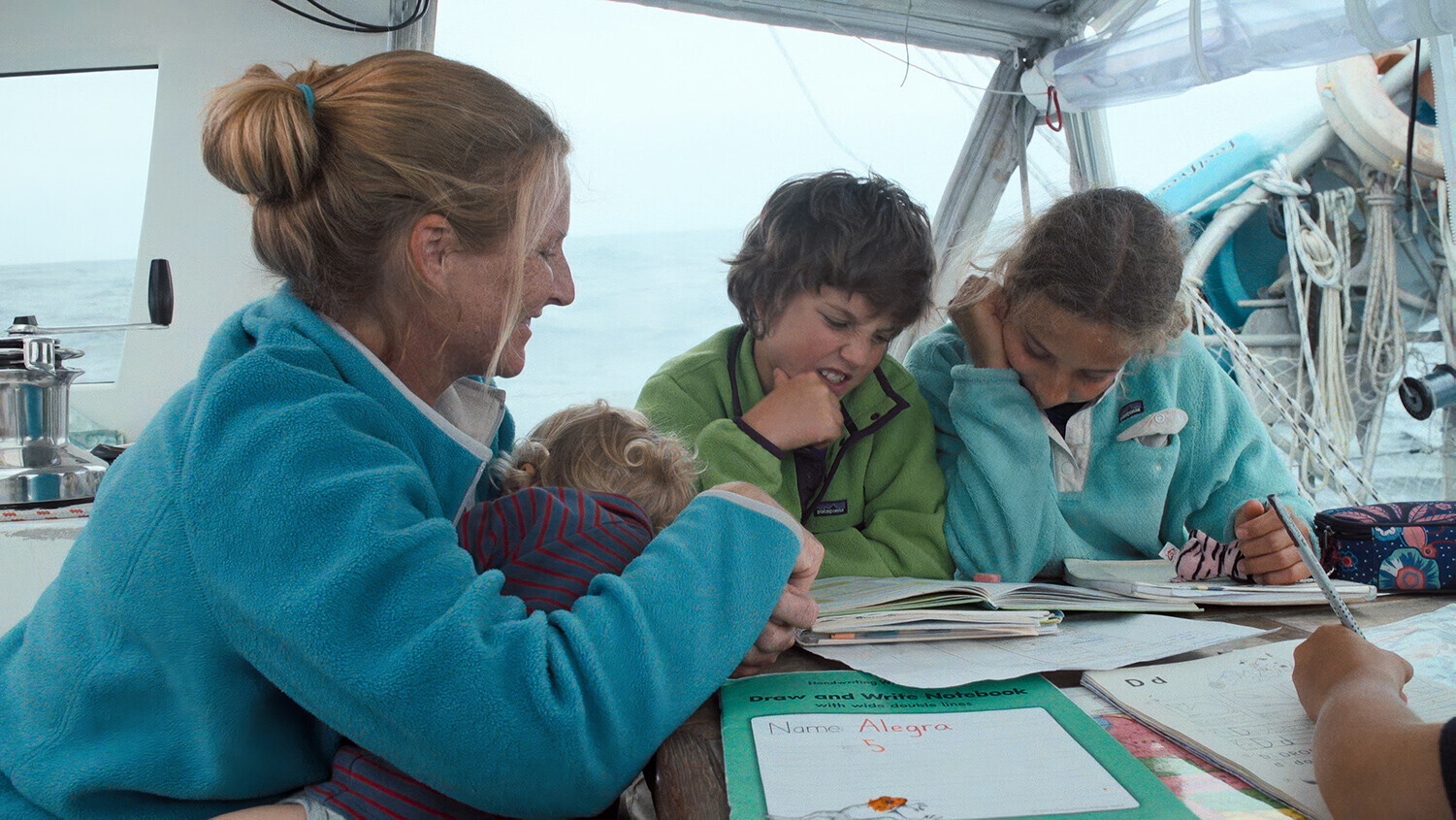 Sabine Schw&ouml;rer unterrichtet drei Kinder auf dem Deck des TOPtoTOP Expeditionssegelbootes, w&auml;hrend sie ein Baby h&auml;lt &ndash; Schulhefte liegen auf dem Tisch